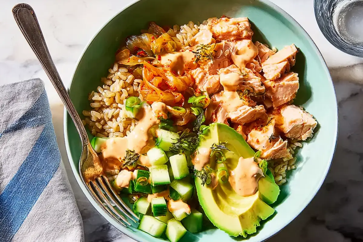 All Recipes Salmon rice bowl recipe with brown rice, avocado, cucumber, kimchi, and spicy mayo served in a green bowl.